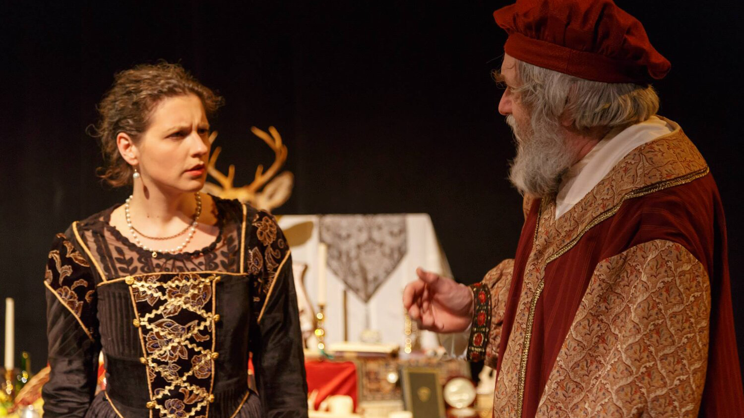 Two actors perform in Richard II. One woman in a black and gold dress looks at a man whose back is to the camera.