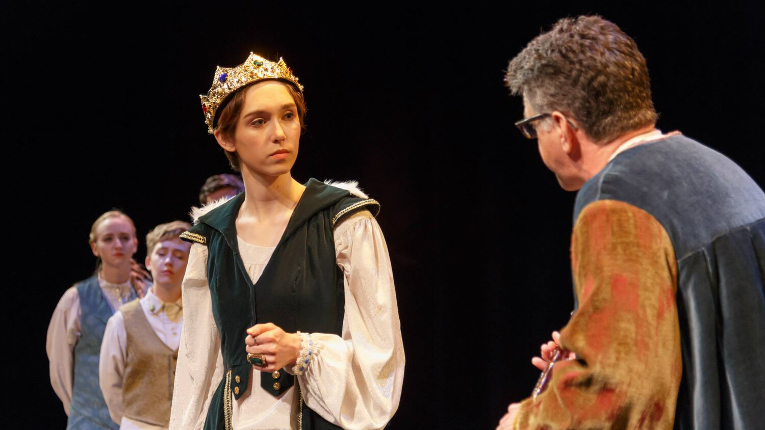 Actors perform in Richard II. One faces the camera wearing a crown.