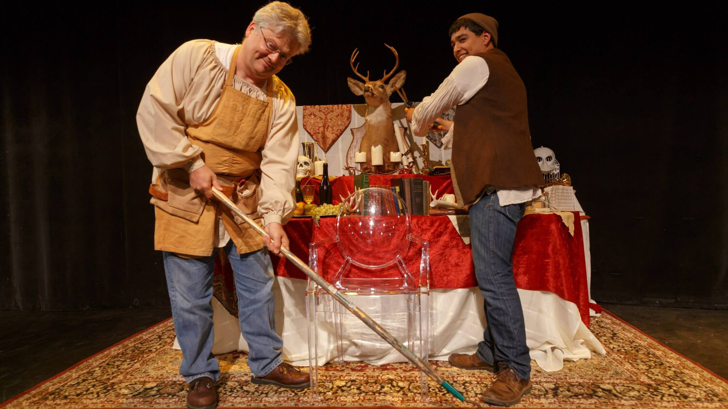 Actors playing gravediggers in Richard II standing in front of a table and clear plastic chair. One is holding a gardening tool.